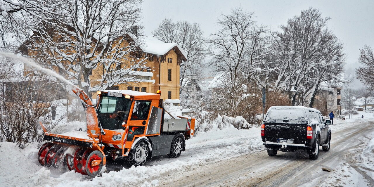 Ein orangefarbener Schneepflug räumt eine verschneite Straße in einem Dorf. Im Hintergrund stehen schneebedeckte Häuser und Bäume. Ein schwarzes Auto fährt auf der geräumten Straße an dem Schneepflug vorbei. Die Szene zeigt winterliche Verhältnisse mit vi