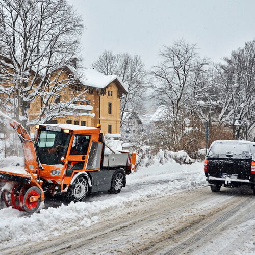 Ein orangefarbener Schneepflug räumt eine verschneite Straße in einem Dorf. Im Hintergrund stehen schneebedeckte Häuser und Bäume. Ein schwarzes Auto fährt auf der geräumten Straße an dem Schneepflug vorbei. Die Szene zeigt winterliche Verhältnisse mit vi