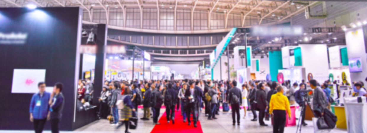 Große Menschenmenge besucht eine lebhafte Messehalle mit verschiedenen Messeständen, bunten Displays und rotem Teppich.